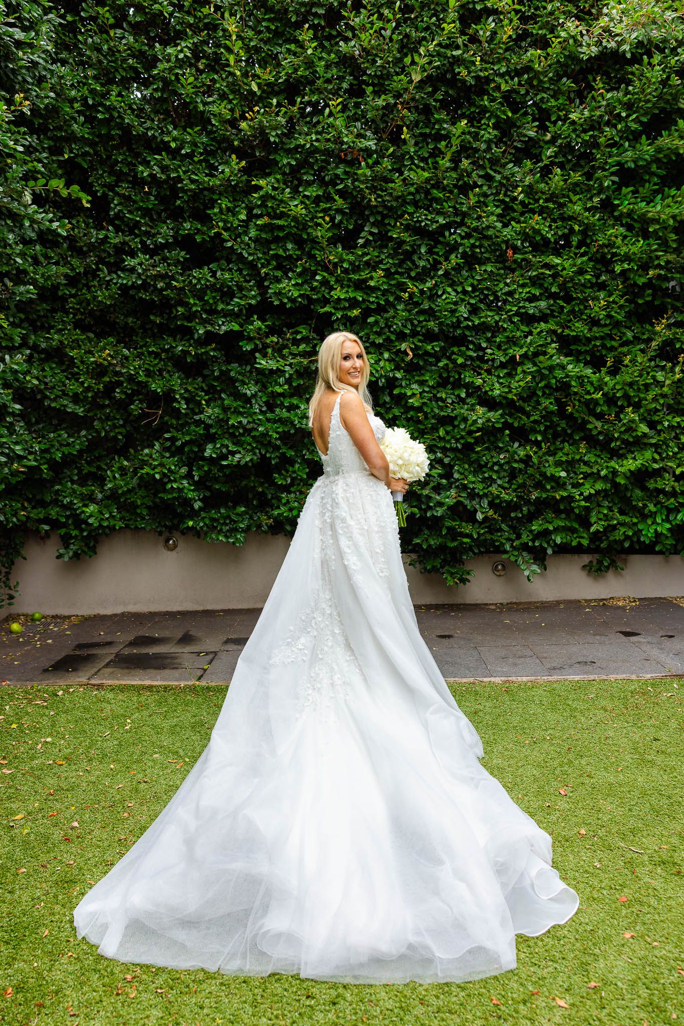 A bride in a flowing white ballgown twirls gracefully on a green lawn with lush hedges in the background.