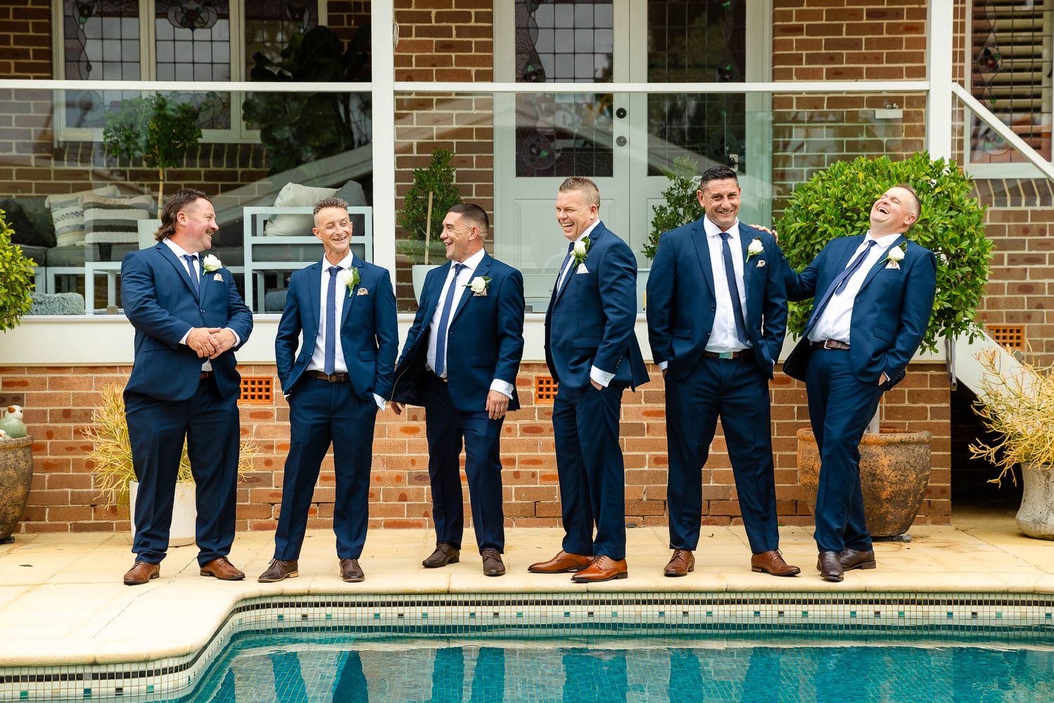 A group in matching navy suits stands casually along the edge of a swimming pool near a brick building.