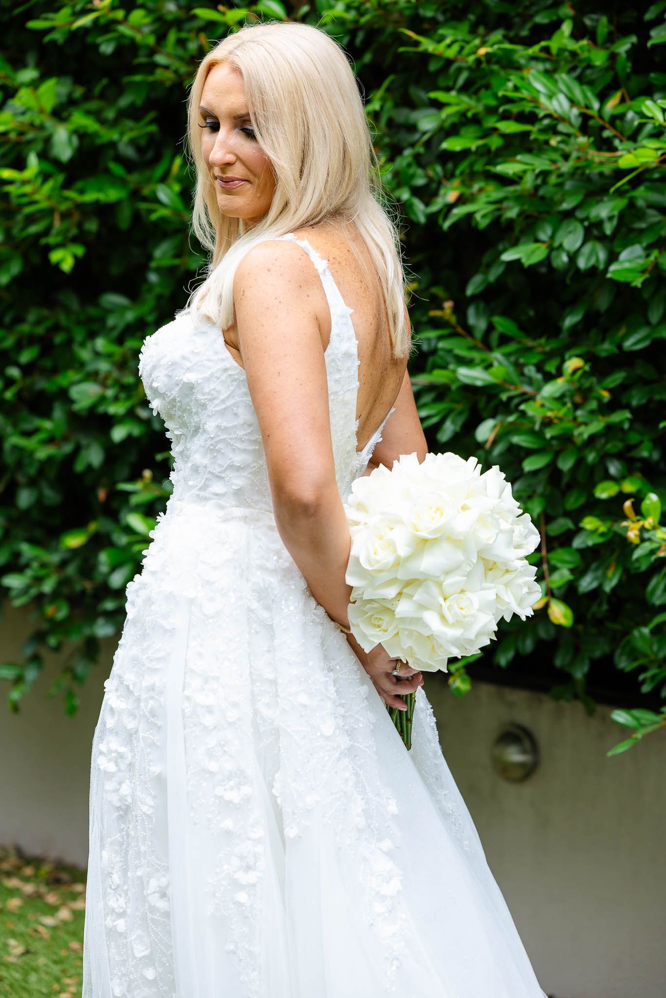 A bride in a white lace wedding dress poses in a garden setting with a large white peony bouquet.