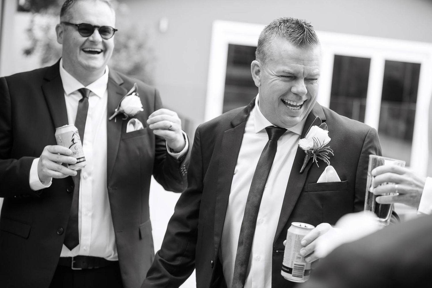 Black and white photo of wedding guests in suits and sunglasses laughing and holding drinks at outdoor reception.