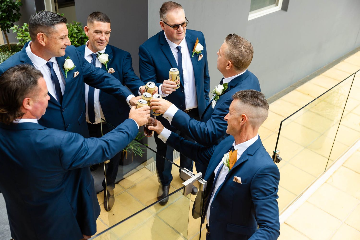Groomsmen in matching navy blue suits toast with beer bottles while standing in a modern venue.