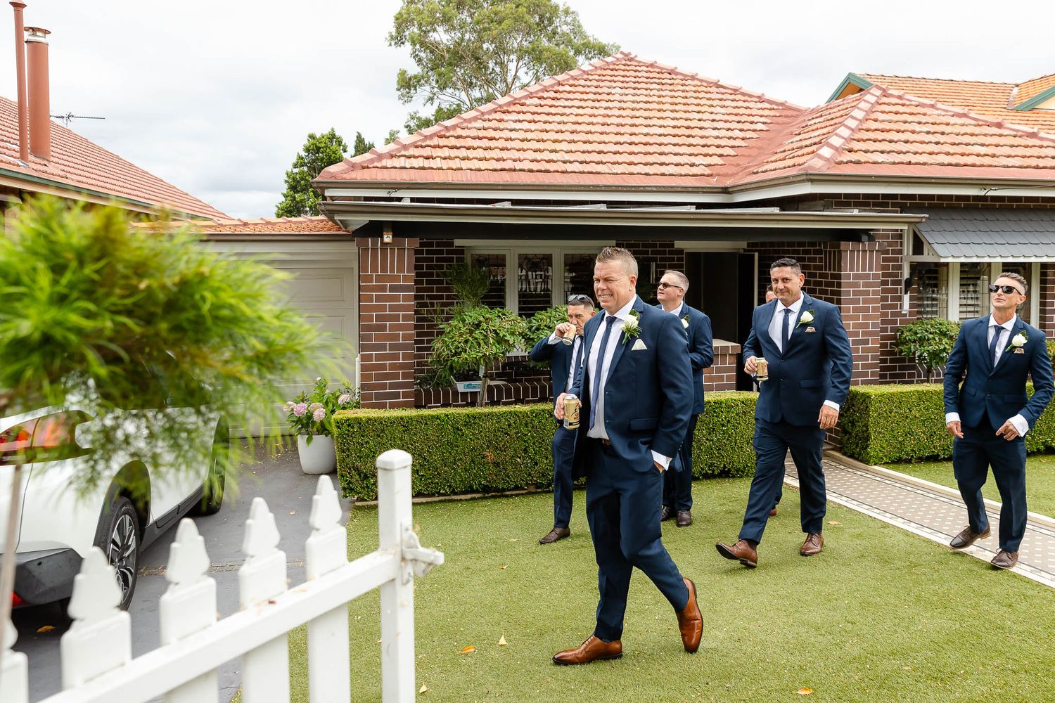 Groomsmen in navy suits walk together on a path outside a building with terracotta roof tiles.