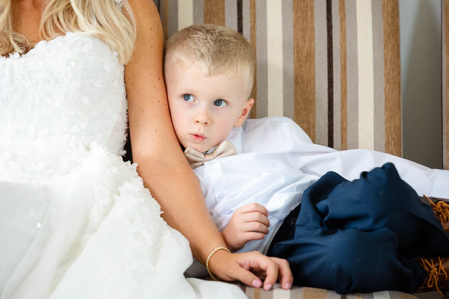 A bride in a white wedding dress sits with a small child wearing formal attire on an elegant sofa.