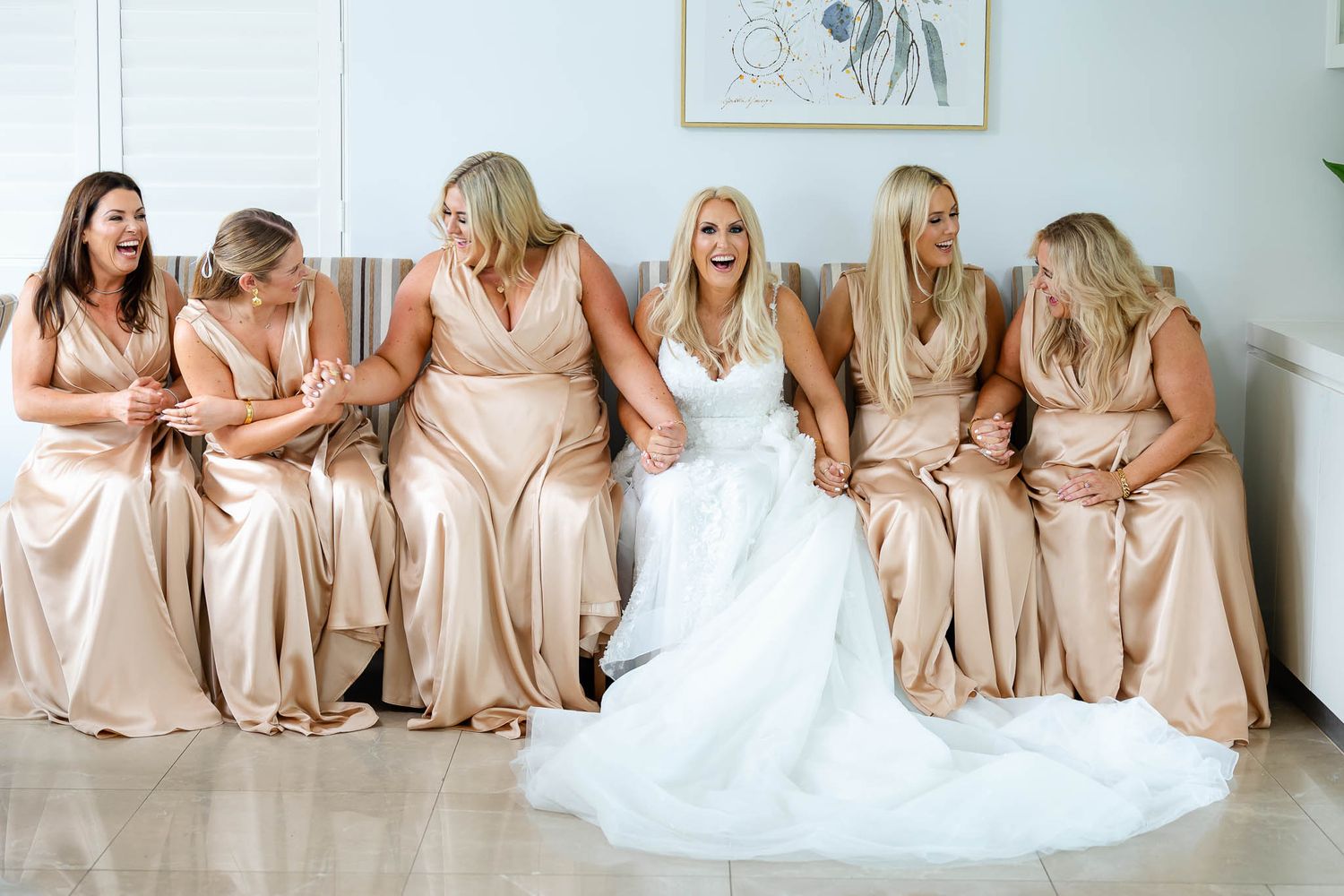 Bridesmaids in champagne colored dresses sit in a row on a white couch with the bride in a white gown.