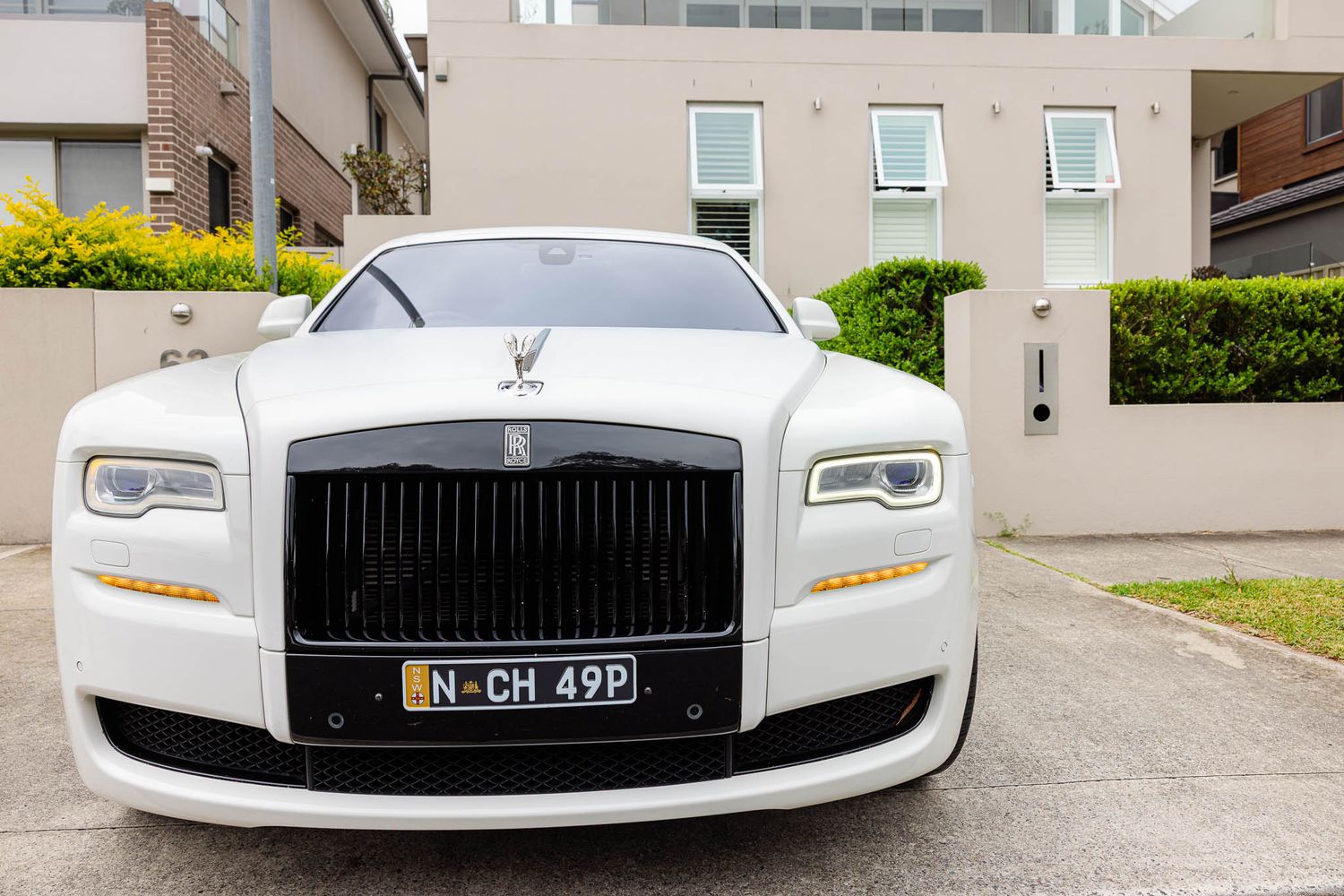 A white Rolls-Royce Ghost luxury sedan with distinctive LED headlights parked in front of modern buildings.