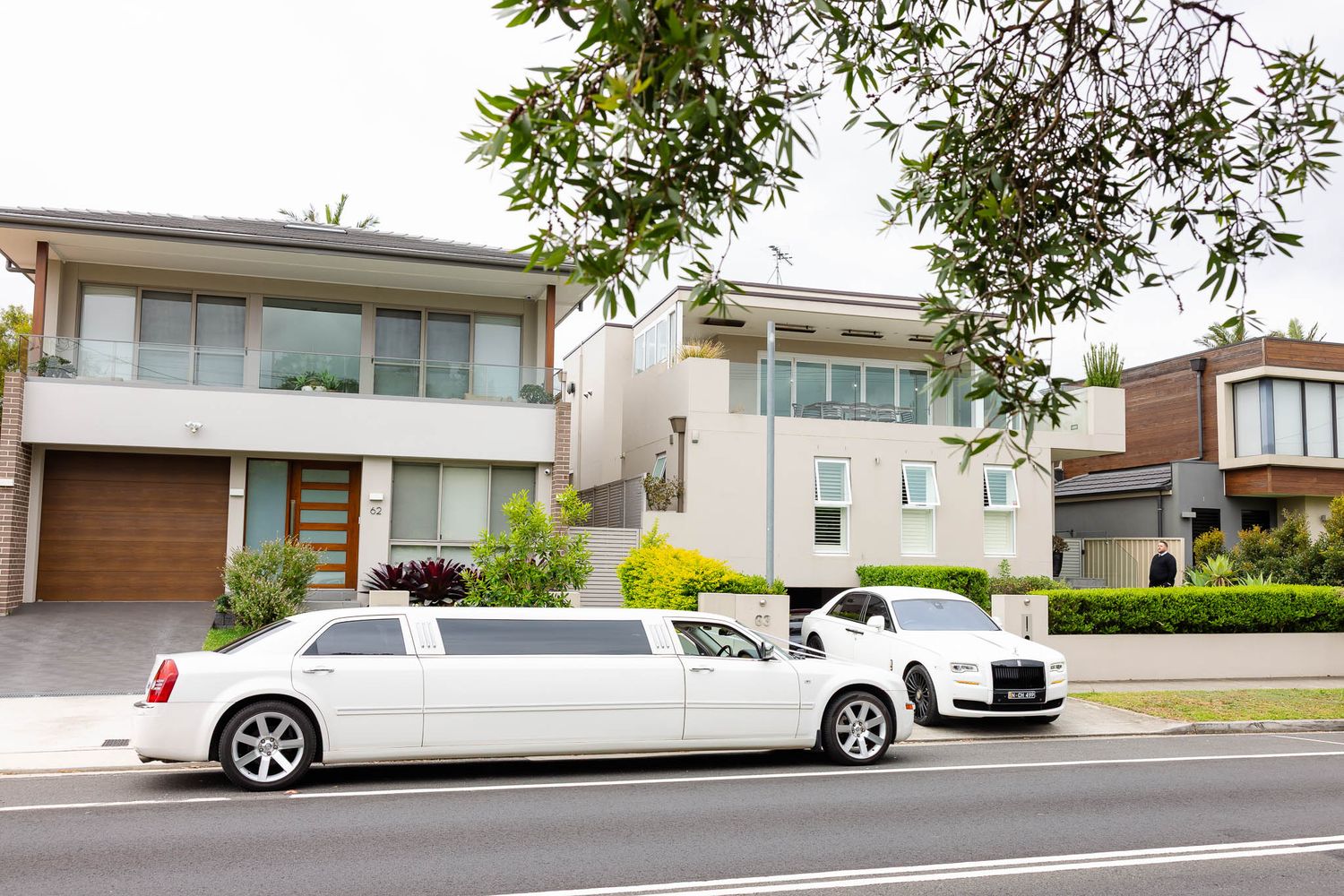 Modern white luxury homes with contemporary architecture line a residential street with parked cars and green trees.