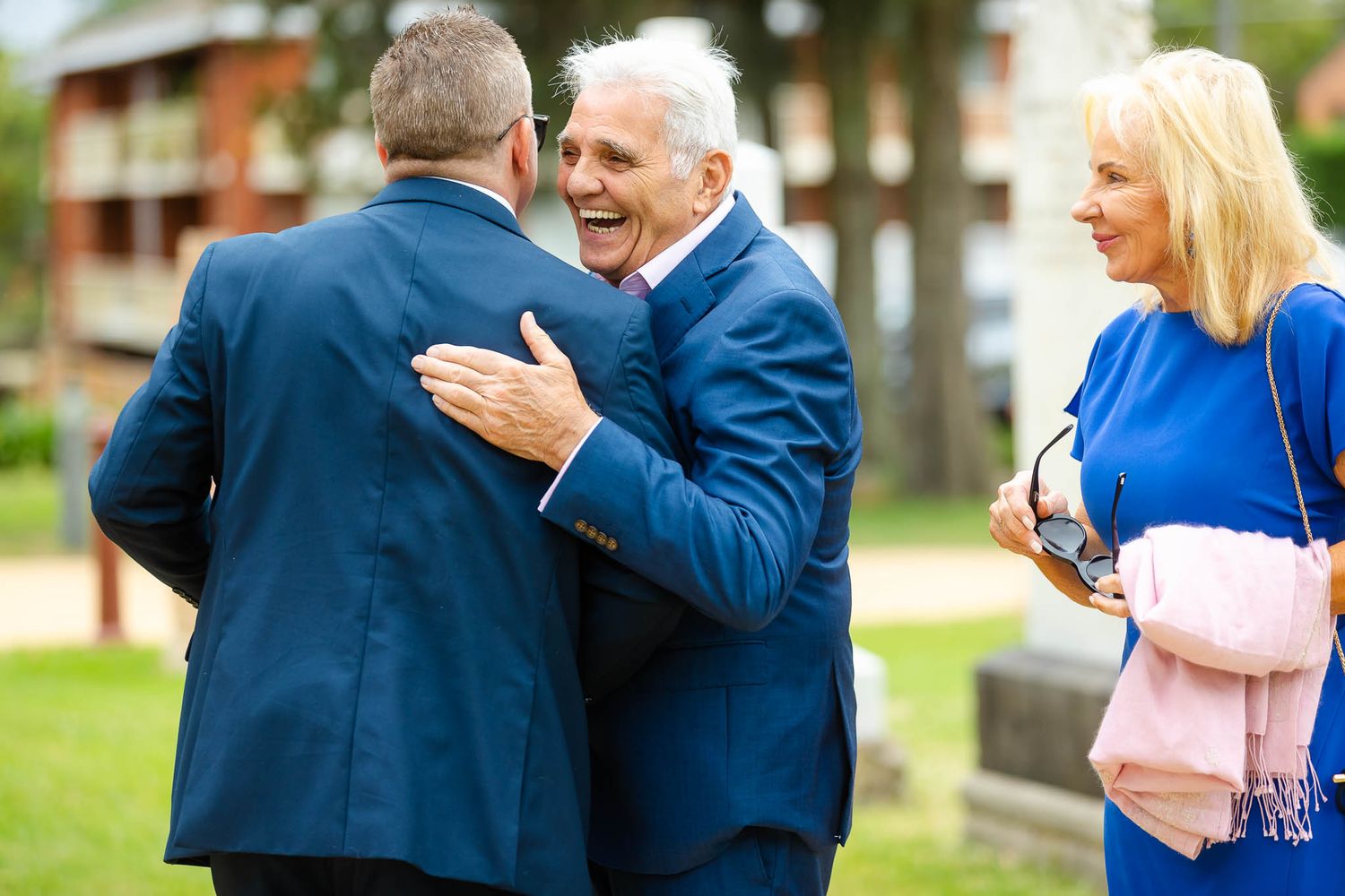 People in blue suits greet each other warmly at an outdoor gathering while a woman in bright blue looks on.