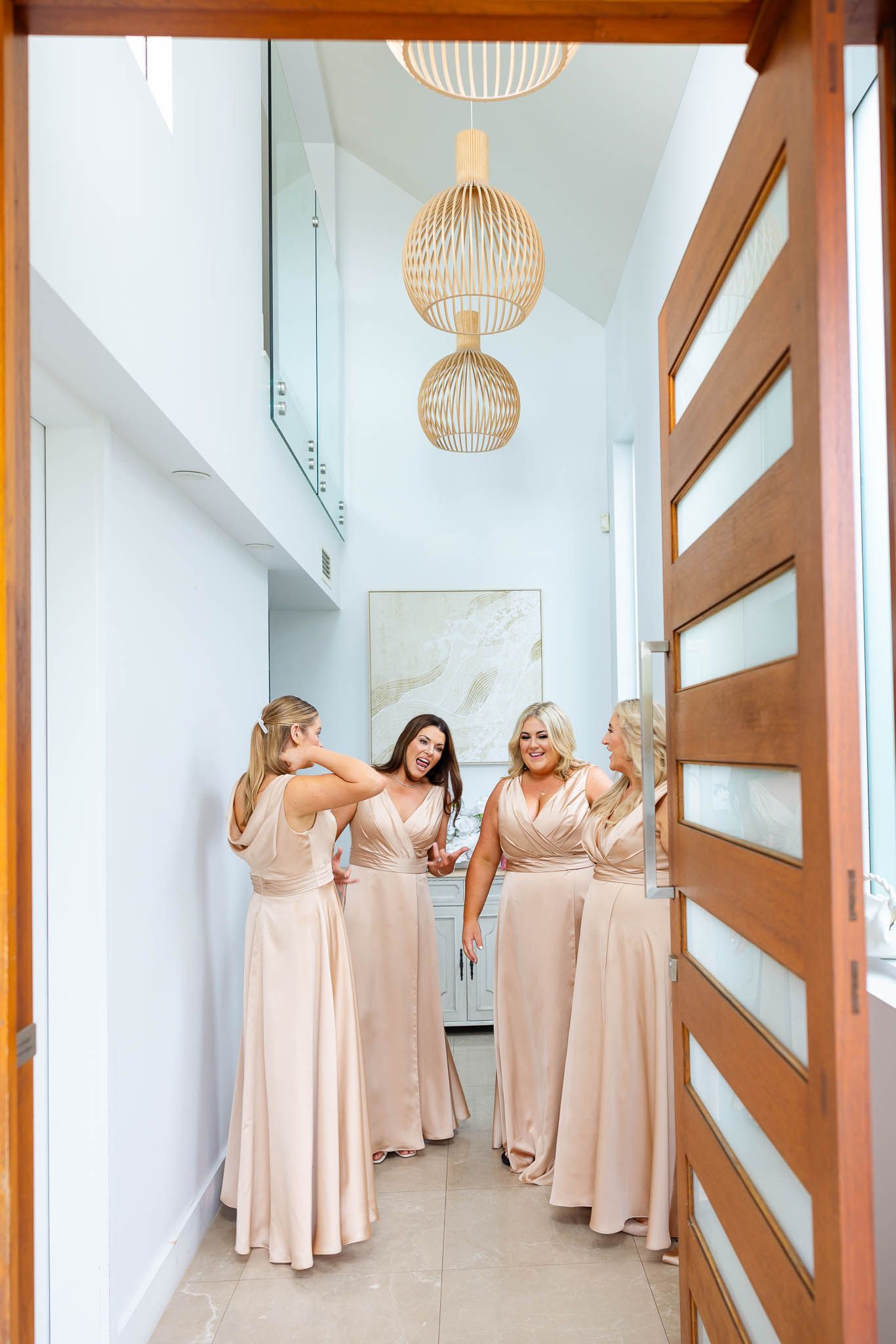 Bridesmaids in blush pink dresses prepare in a modern white hallway with wooden doors and woven pendant lights.