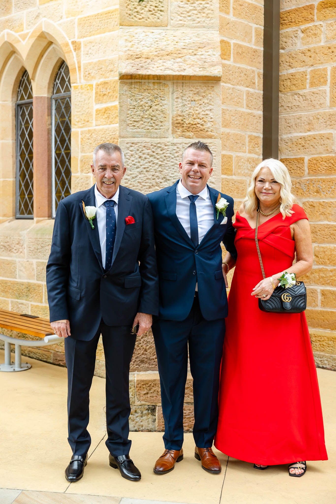 Wedding party members in formal attire with men in black suits and women in bright red dresses pose against stone wall.