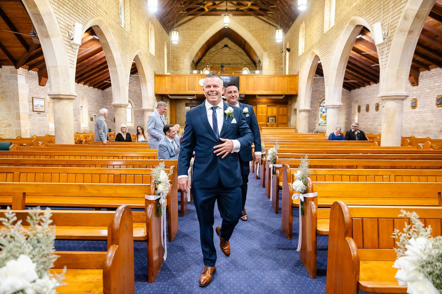 Wedding party members walk down the aisle of a grand cathedral with soaring arches and wooden pews.
