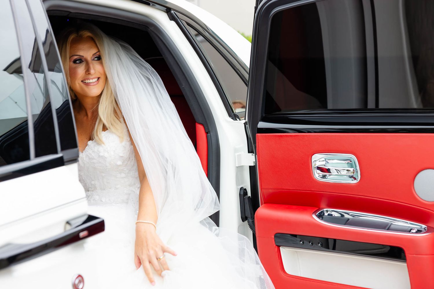 A bride in a white wedding dress enters a red car door while holding her gown.