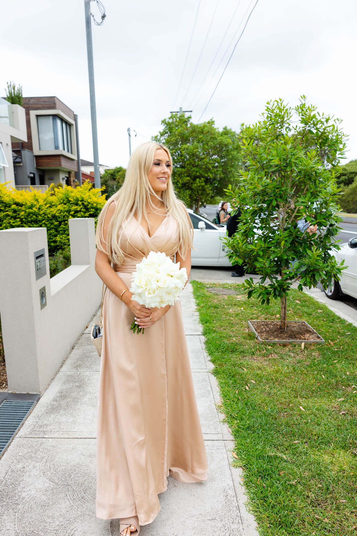 A person in a flowing champagne-colored dress stands on a paved walkway beside a landscaped garden area.