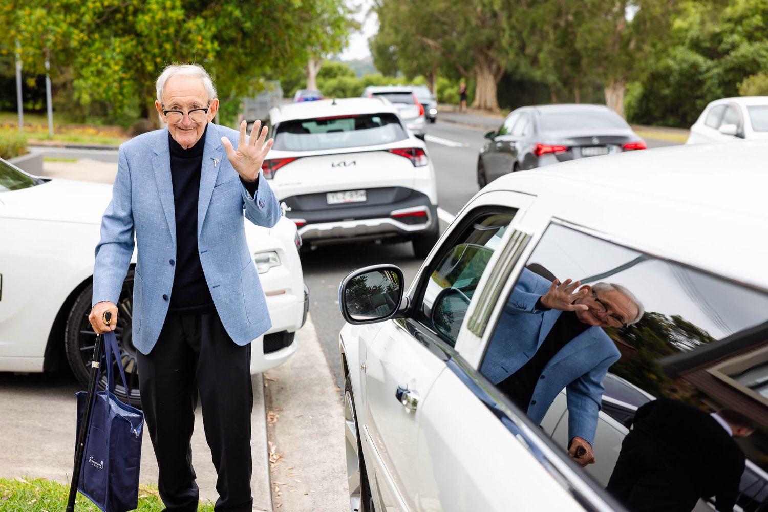 A person in a blue jacket stands next to a parked car in a parking lot on a sunny day.