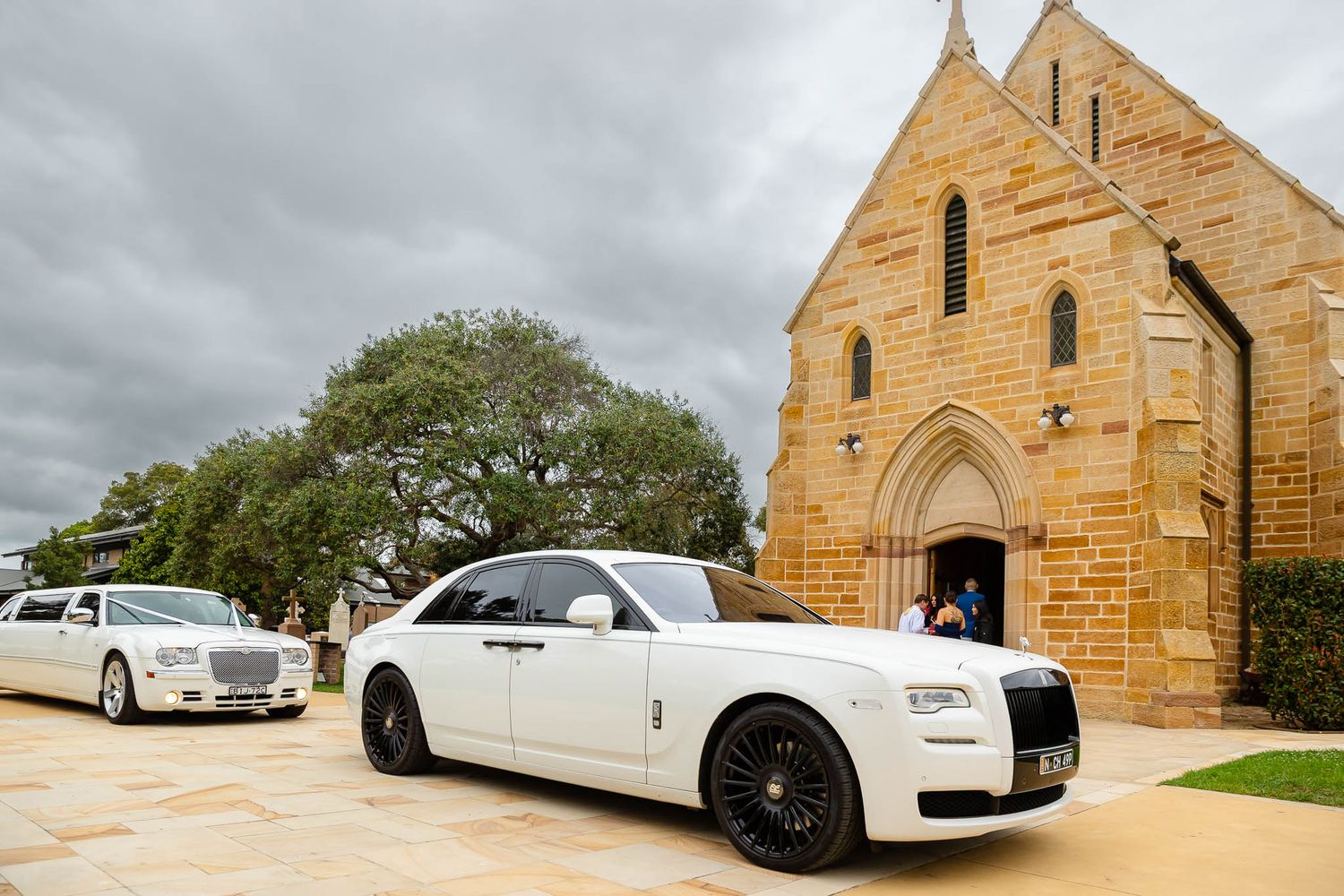 White Rolls Royce luxury wedding car parked in front of a sandstone church on an overcast day.