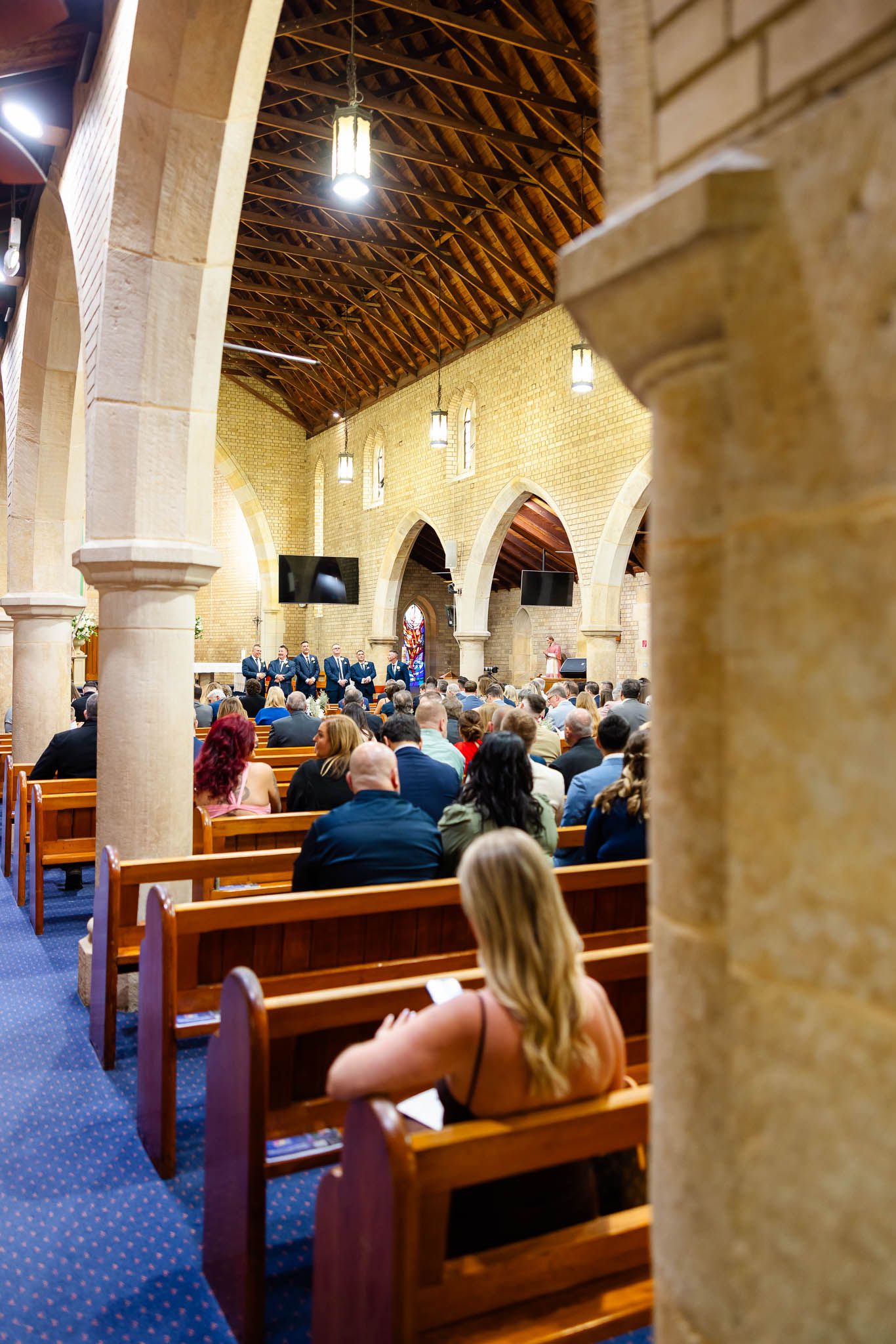 Church congregation seated in wooden pews under vaulted ceiling with stone columns and pendant lights.