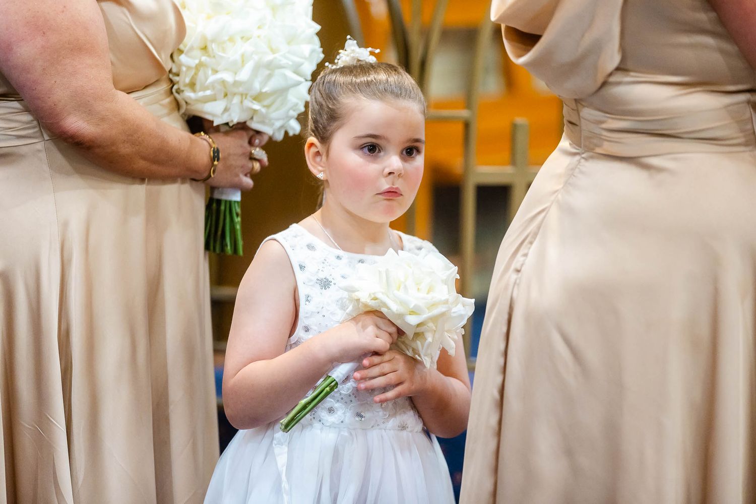 A young flower girl in a white dress holds a bouquet while standing between bridesmaids in beige dresses at a wedding.