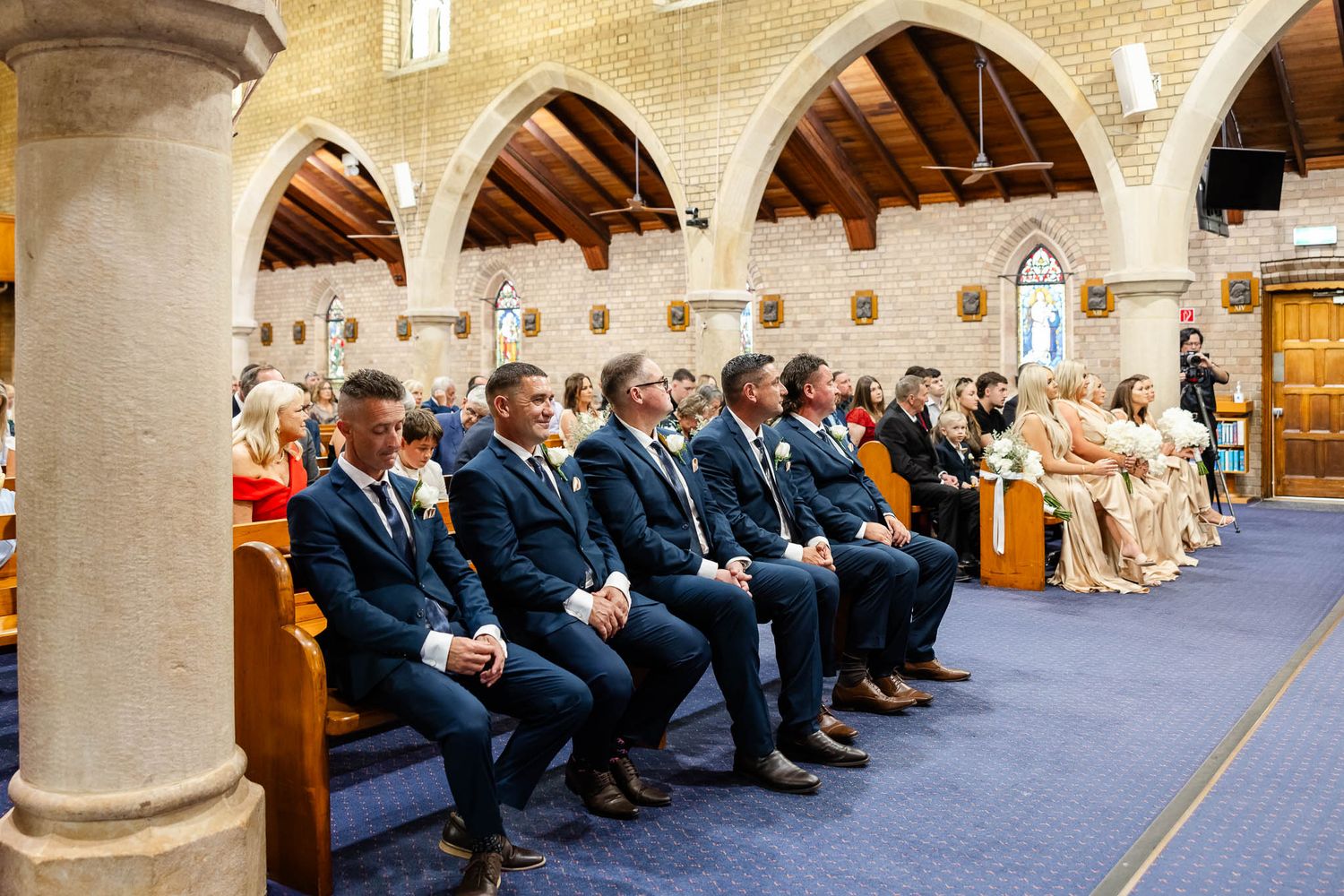 Wedding guests seated in church pews during a ceremony in a stone cathedral with natural lighting.
