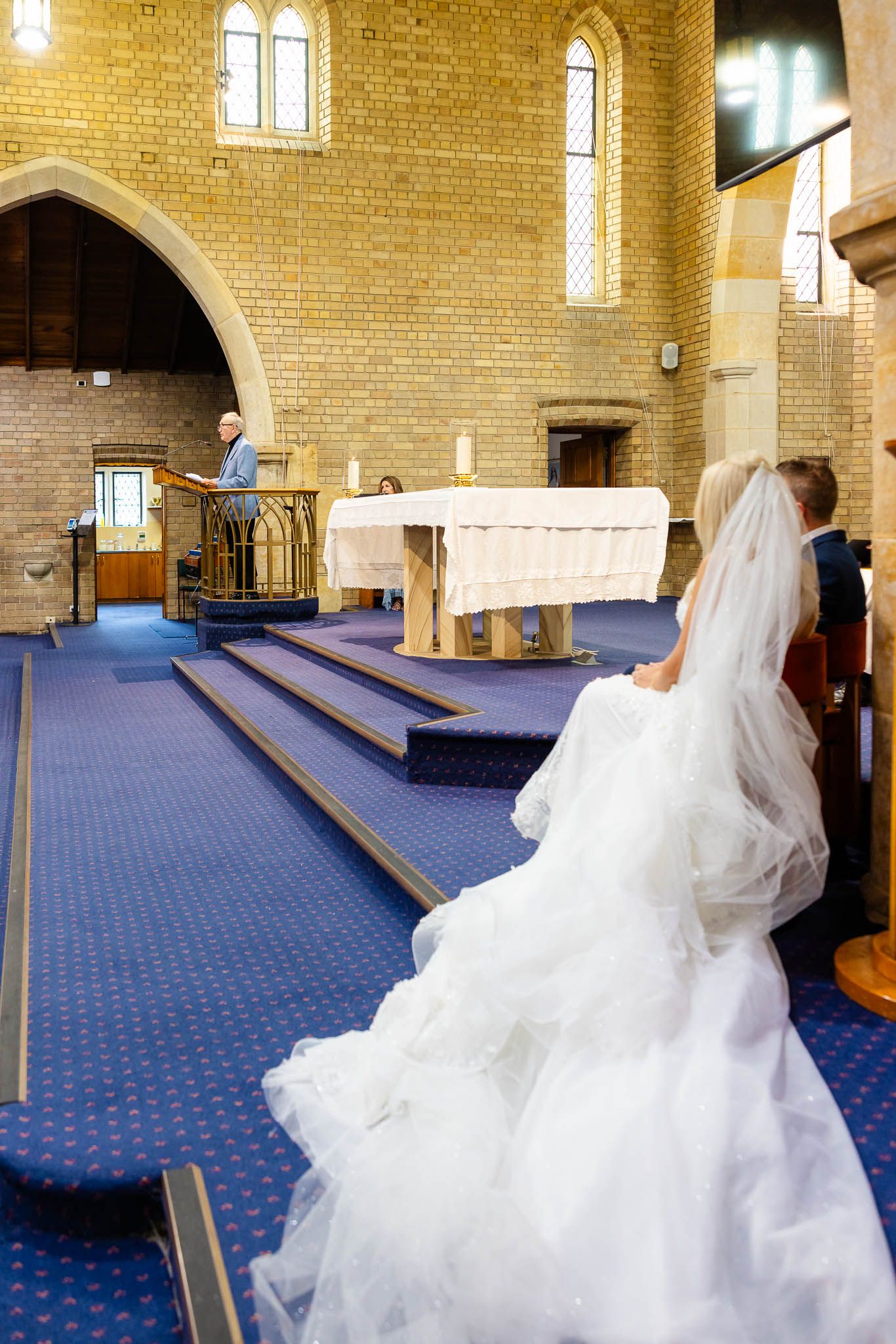 A white wedding dress twirls across a blue carpeted floor inside a brick-walled church sanctuary.