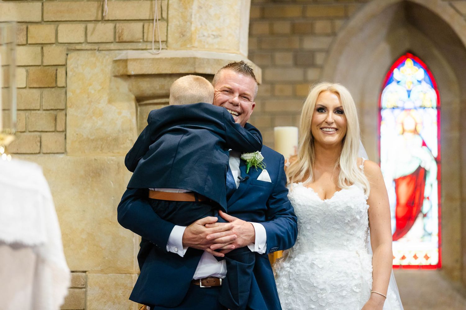 A happy couple embraces and laughs together during their wedding ceremony in a stone church with stained glass windows.