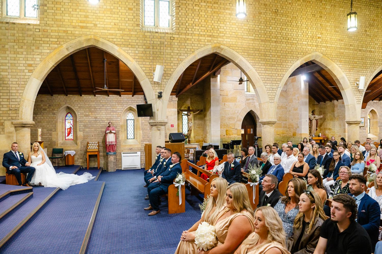 Wedding ceremony taking place inside a historic stone church with arched columns and a blue carpeted aisle.