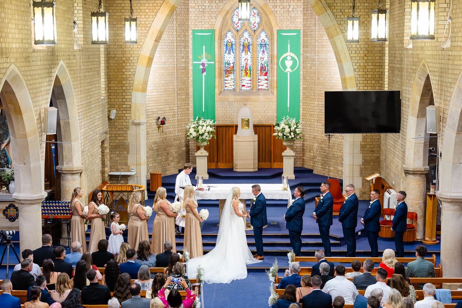 Wedding ceremony taking place in a beautiful stone church with stained glass windows and hanging pendant lights.