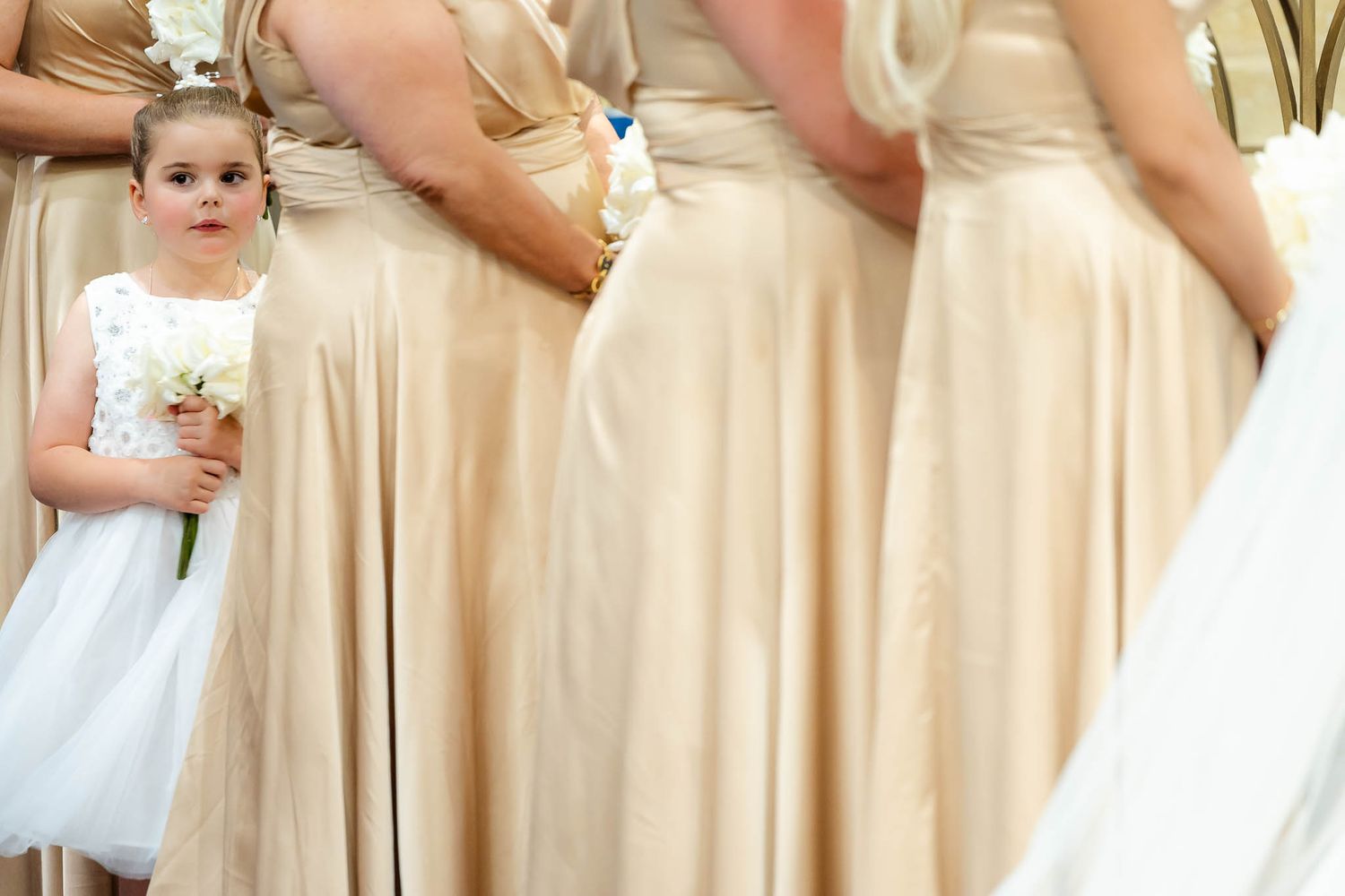 A young attendant in a white tulle dress stands among bridesmaids wearing champagne-colored gowns at a wedding ceremony.