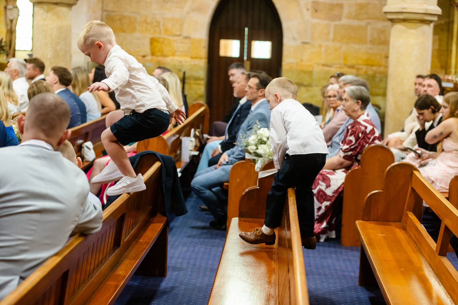 Wedding guests seated in wooden church pews watch a ceremony unfold in a historic stone chapel.