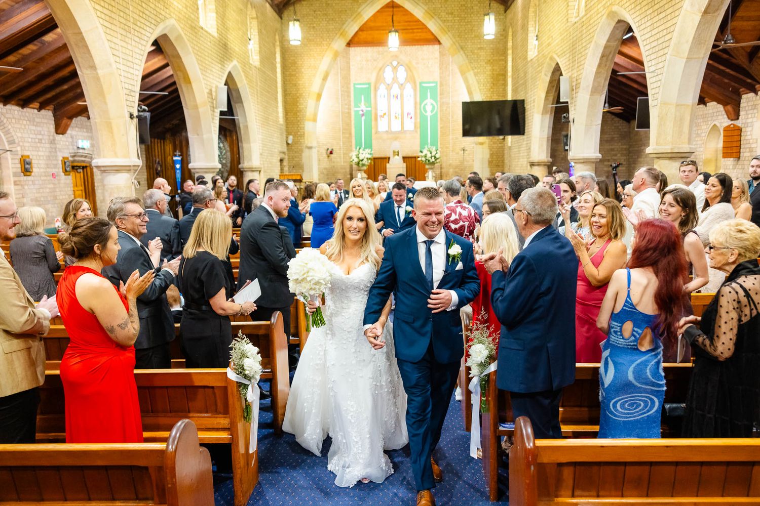 Wedding guests stand in church pews during a traditional Catholic ceremony while a couple exchanges vows at the altar.