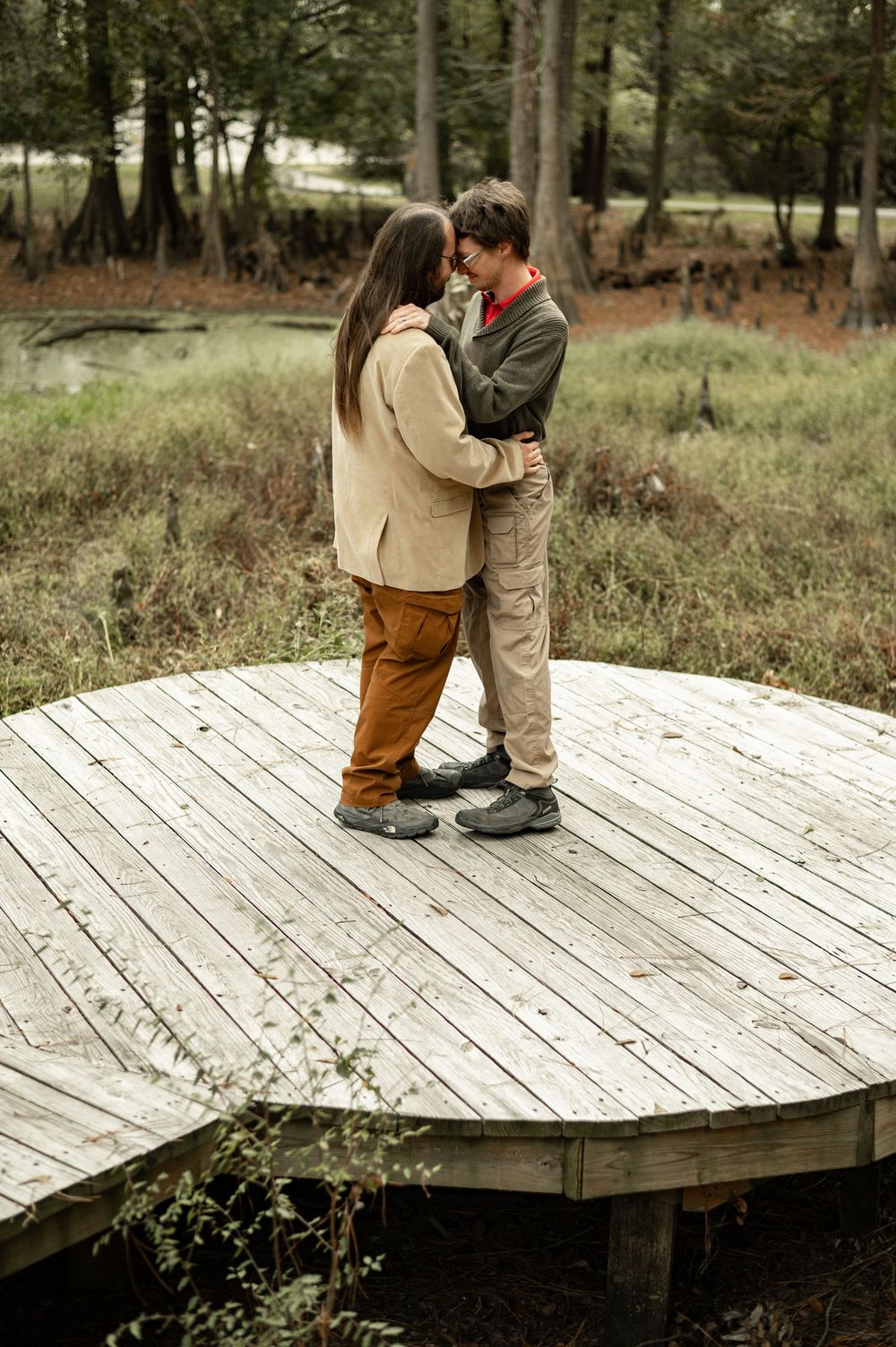 A couple stands intimately together on a rustic wooden deck platform in a wooded outdoor setting.