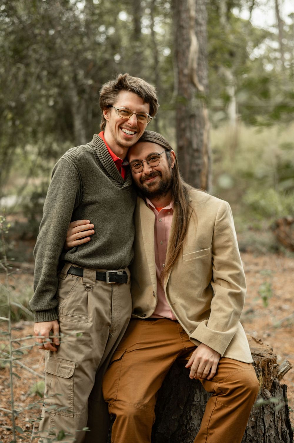 Two people in autumn attire share a tender moment while sitting on a tree stump in a wooded forest setting.