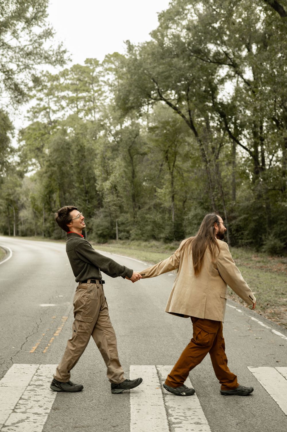 Two people hold hands while walking across a crosswalk on a tree-lined road in casual autumn attire.