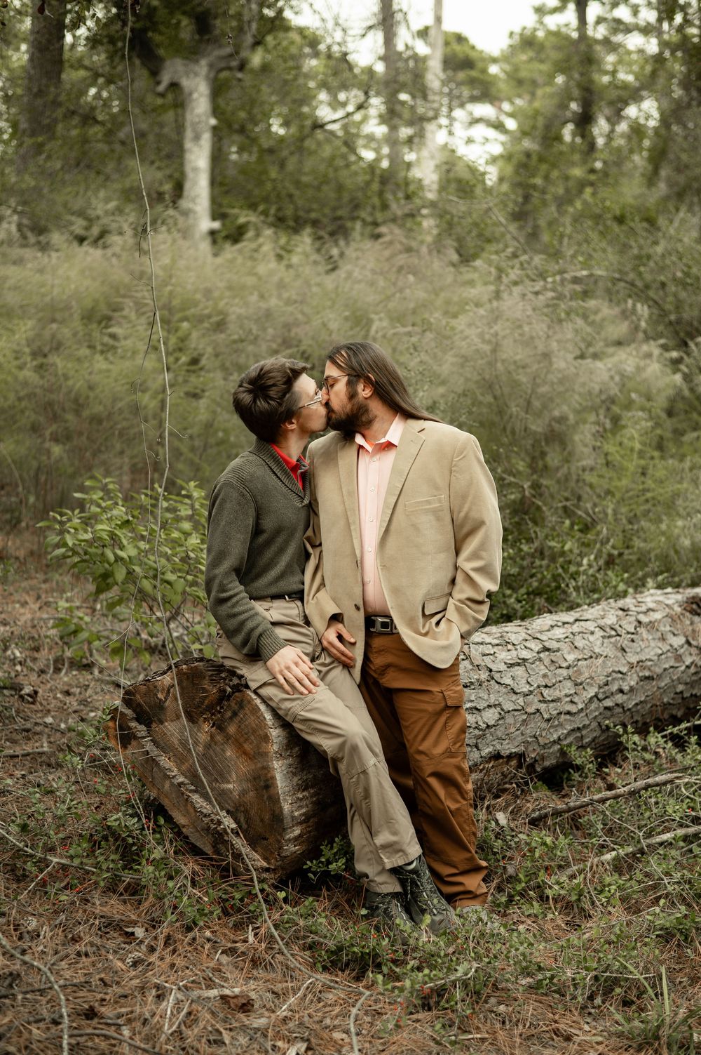 Two people share an intimate moment while sitting on a fallen log in a wooded forest setting.