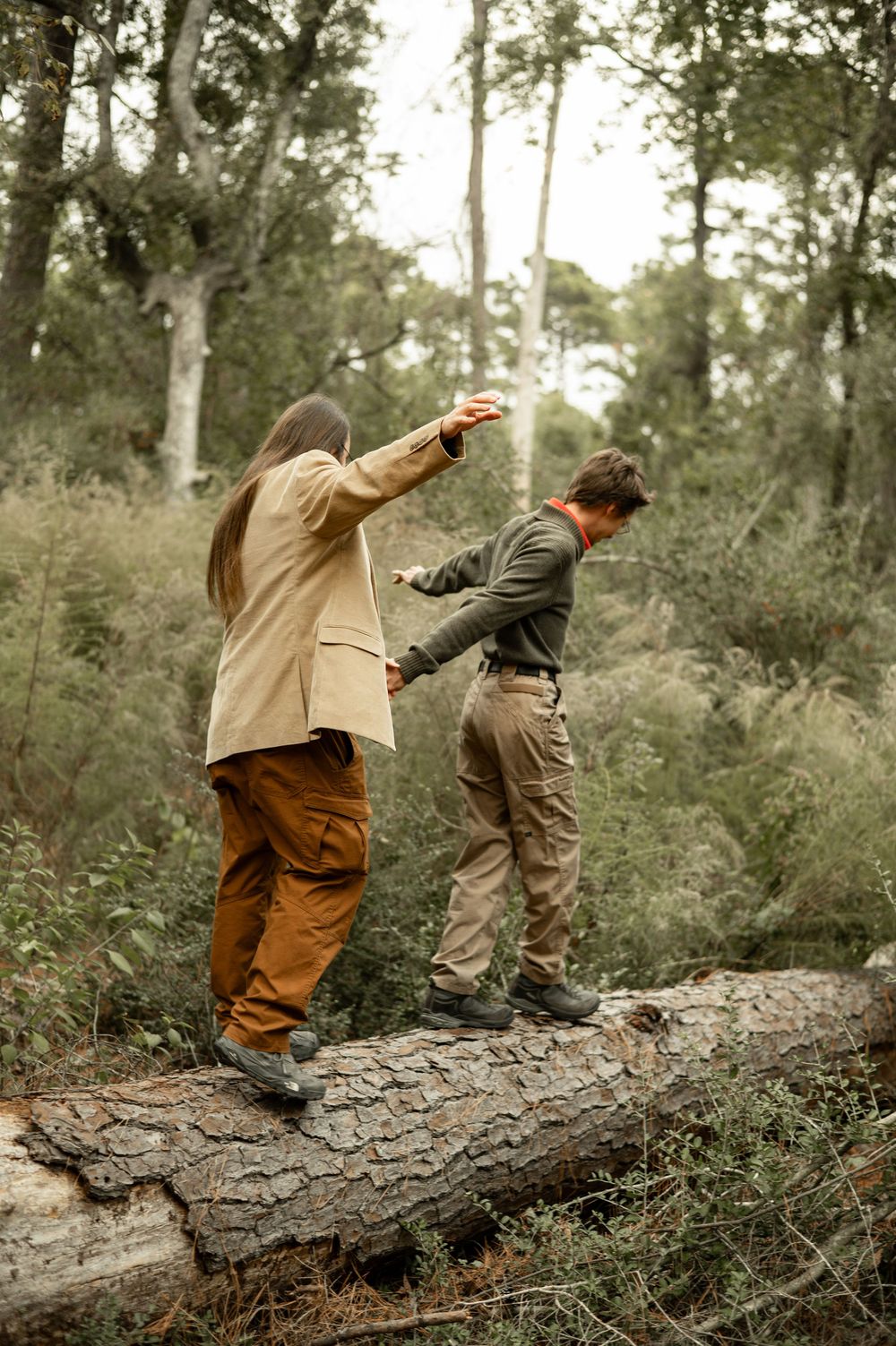 Two hunters in outdoor gear aim their shotguns while standing on a fallen log in a wooded area.