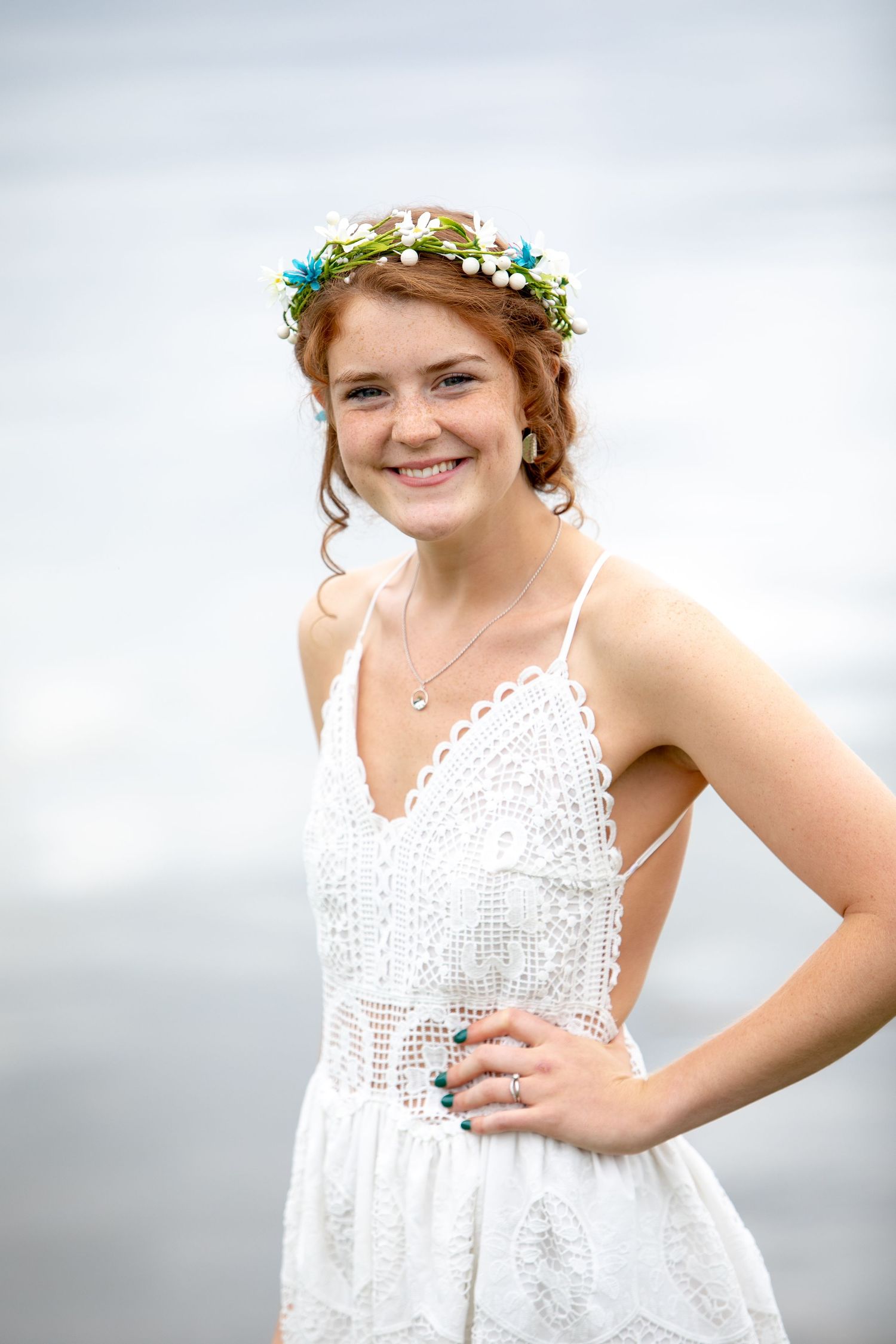 A person wearing a white lace sundress and floral crown poses against a cloudy sky background.