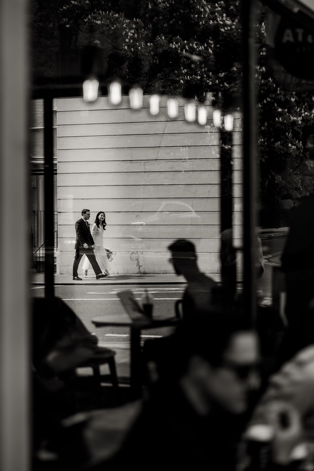 Black and white image showing silhouettes walking along an illuminated wall with hanging lights.