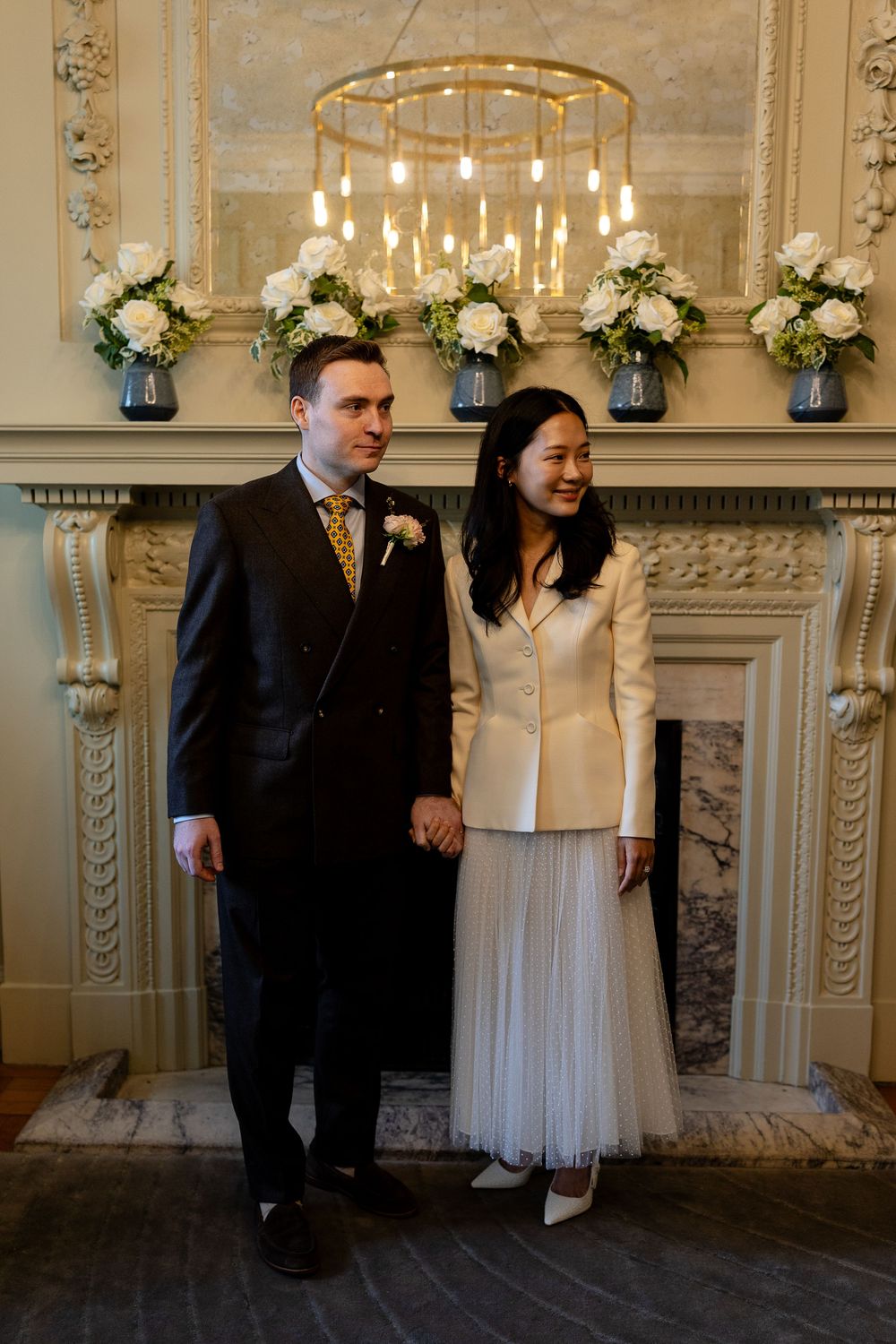 Elegant wedding couple stands in front of ornate fireplace with chandelier and white floral arrangements.