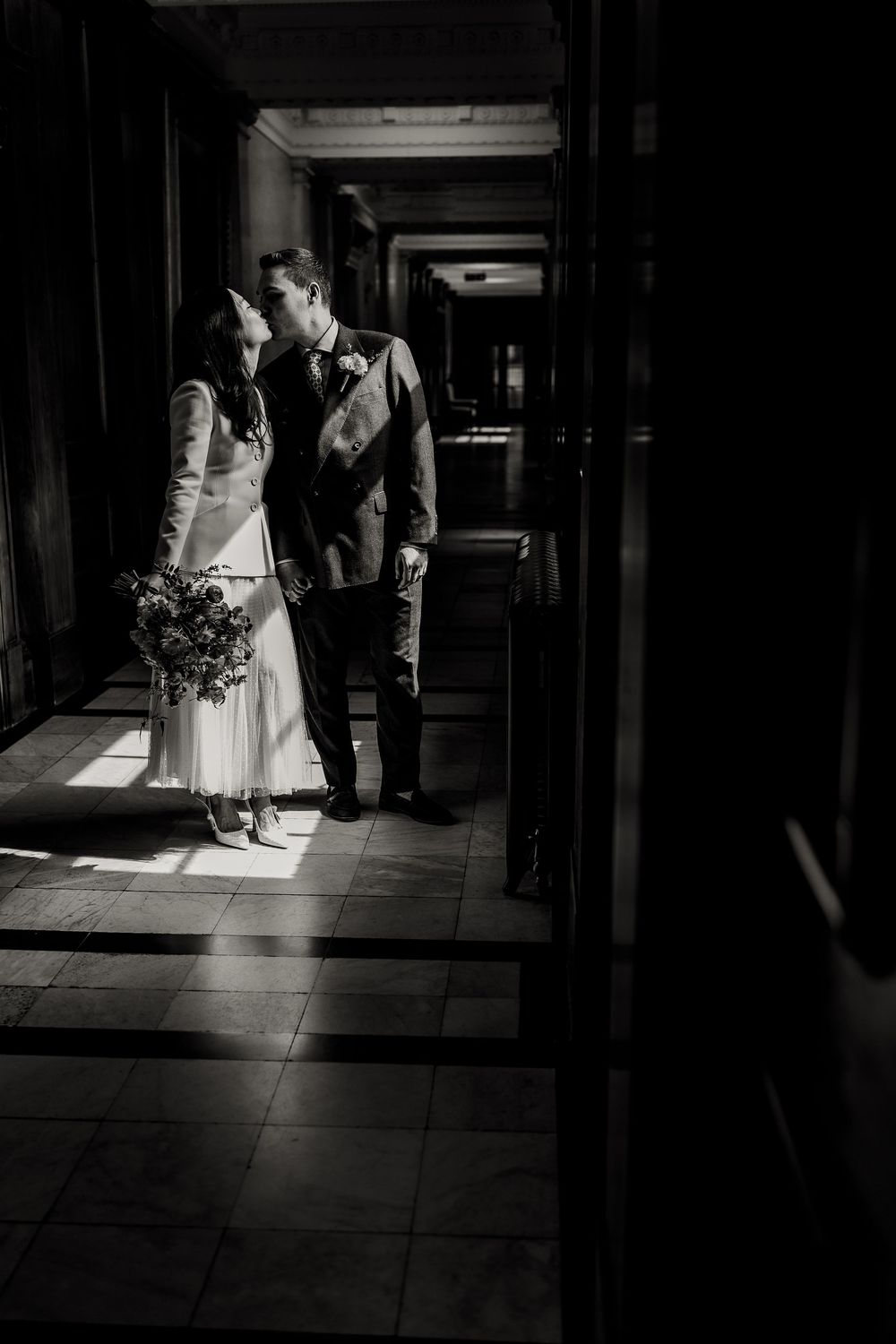 A romantic black and white wedding photo of a couple embracing in a sunlit hallway with dramatic shadows.