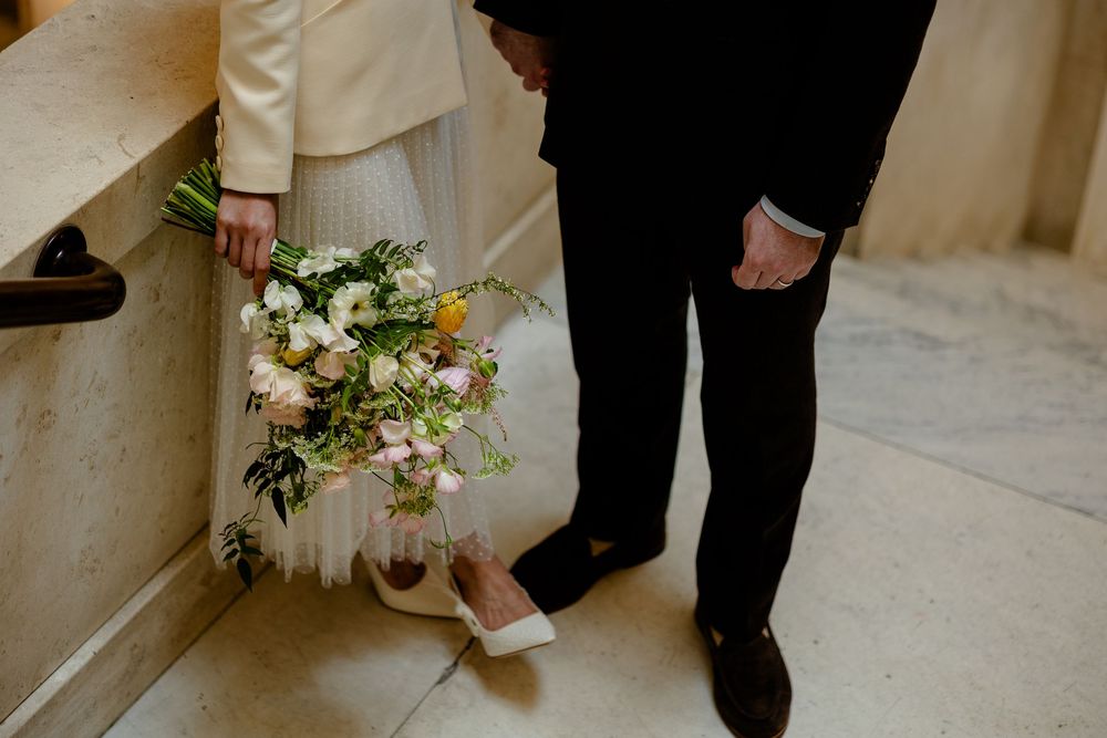 Close-up view of a wedding bouquet with white and peach flowers held next to formal attire and dress shoes.