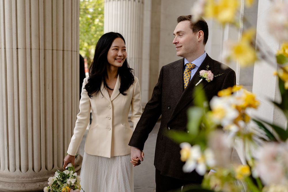 A couple shares a joyful moment among white columns and spring flowers during a wedding photoshoot.