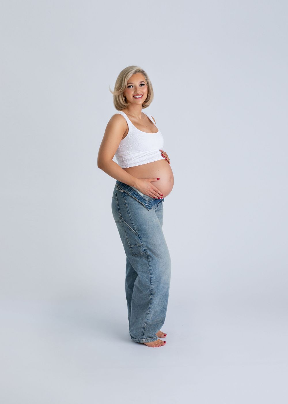 Expectant mother in white tank top and jeans poses sideways showing pregnant belly against light gray background.