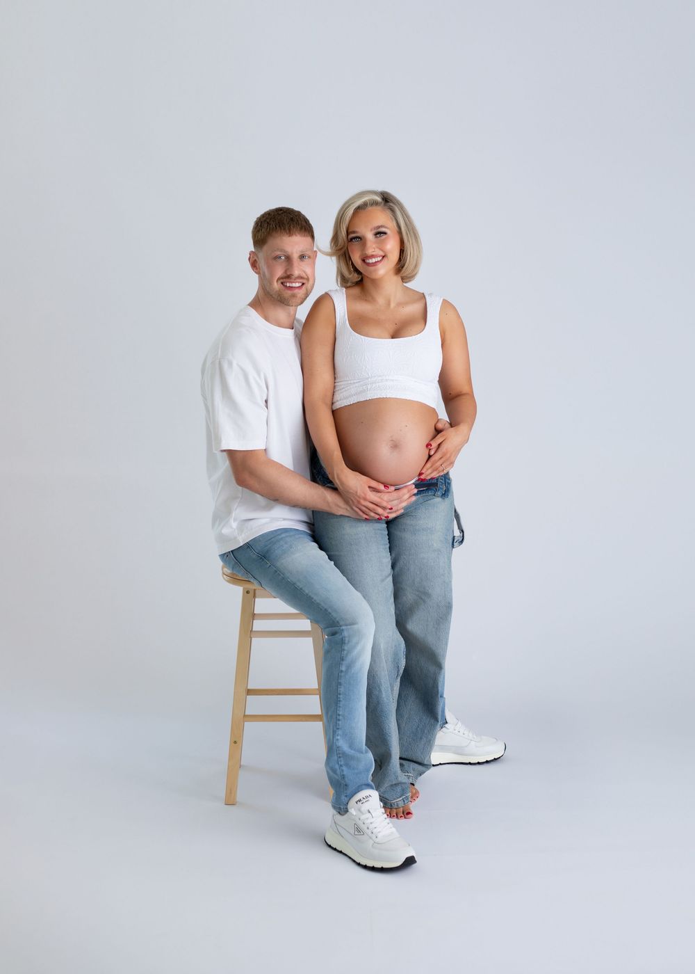 A couple sits together on a wooden stool against a white backdrop in a casual, intimate photoshoot.