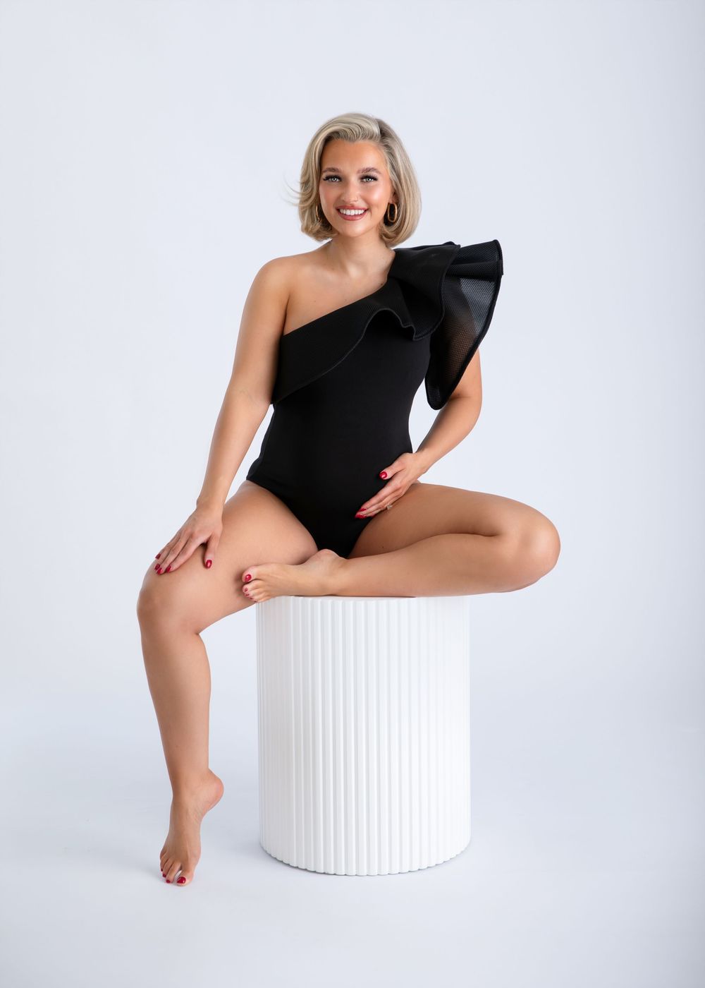 A figure in a black one-shoulder dress poses elegantly on a white cylindrical ottoman against a light background.