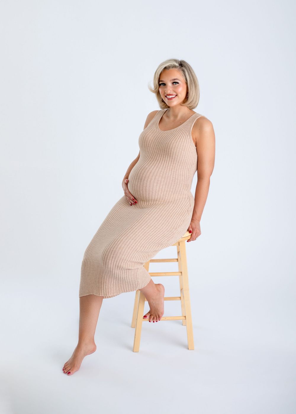 Person in beige ribbed sweater dress poses playfully on wooden stool against white background in photo series.