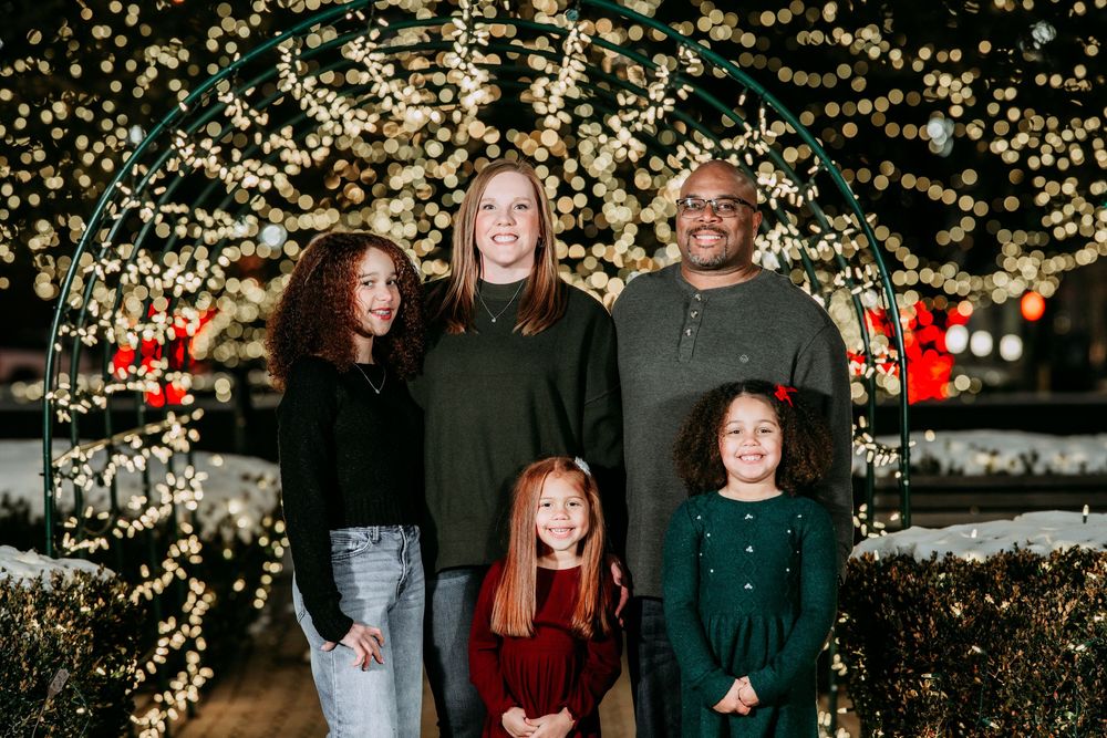 Family poses together outdoors at night with glowing Christmas lights creating a magical holiday backdrop.