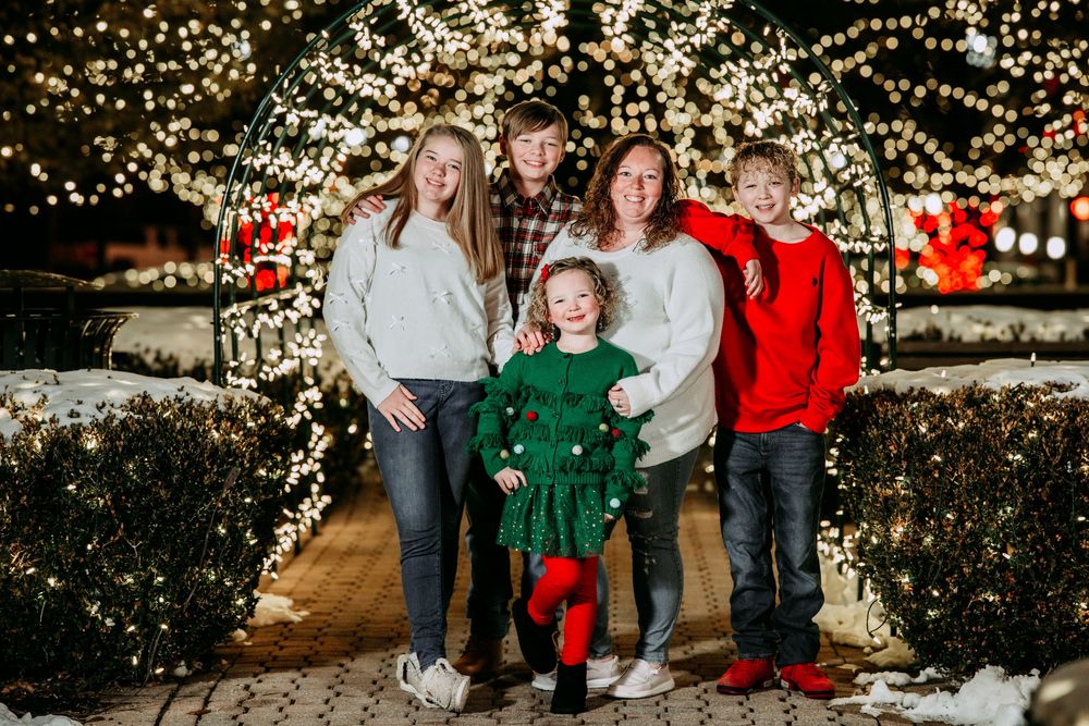 A group poses for holiday photos outdoors at night surrounded by twinkling Christmas lights and snowy scenery.