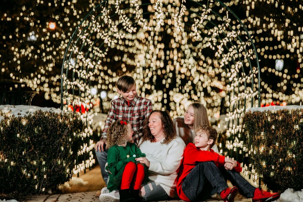 Family wearing festive holiday outfits poses in front of a Christmas tree with glowing lights in the background.