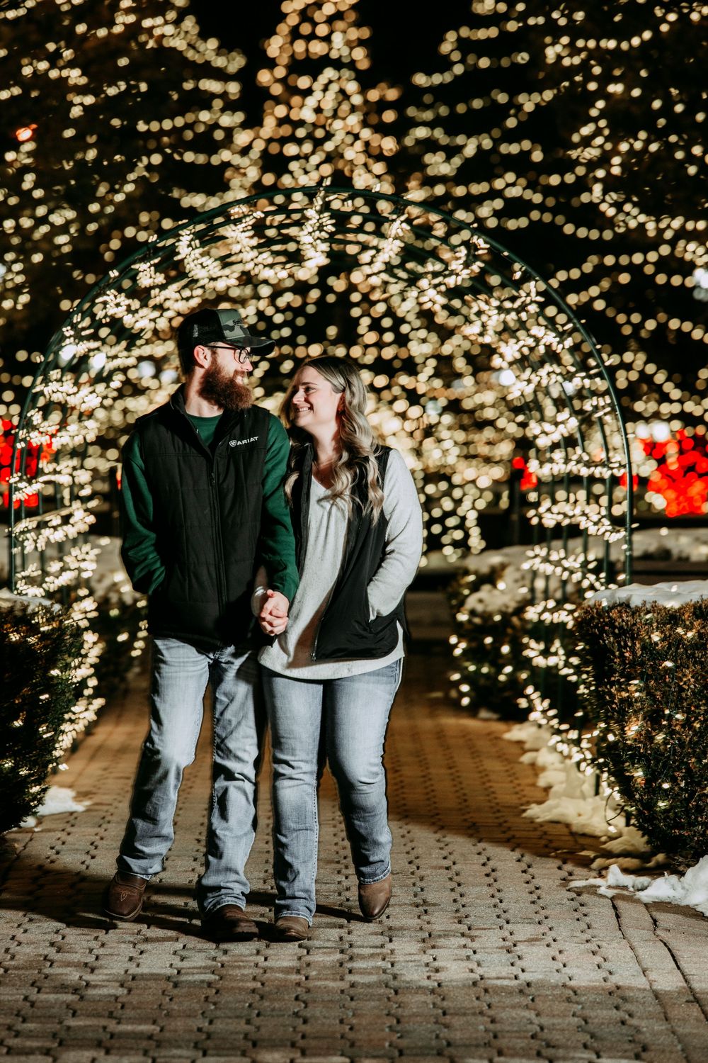 Couple walking together on brick pathway at night surrounded by twinkling holiday lights and illuminated trees.