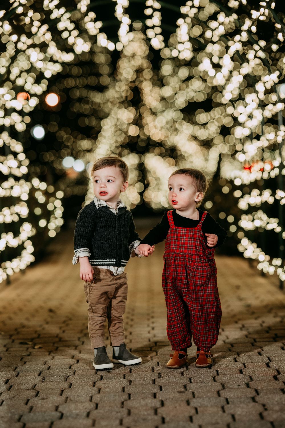 Two young children stand together on a brick path surrounded by magical holiday lights creating a glowing tunnel effect.