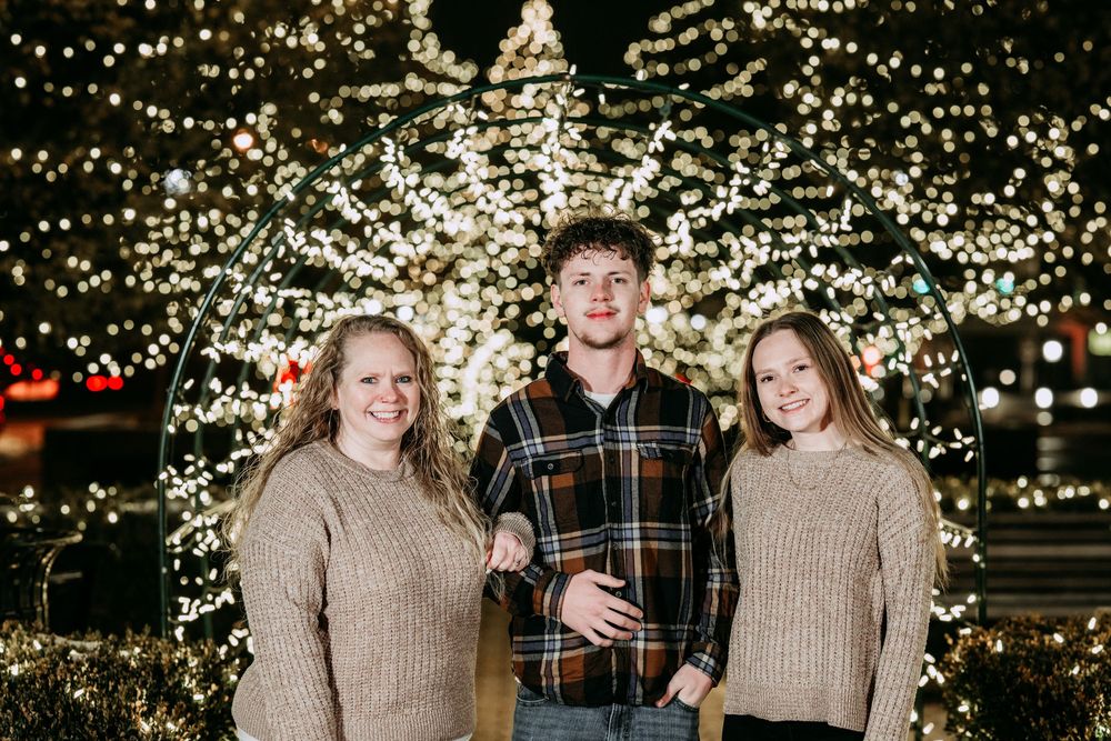 Three people in sweaters stand together smiling in front of illuminated trees with twinkling Christmas lights at night.