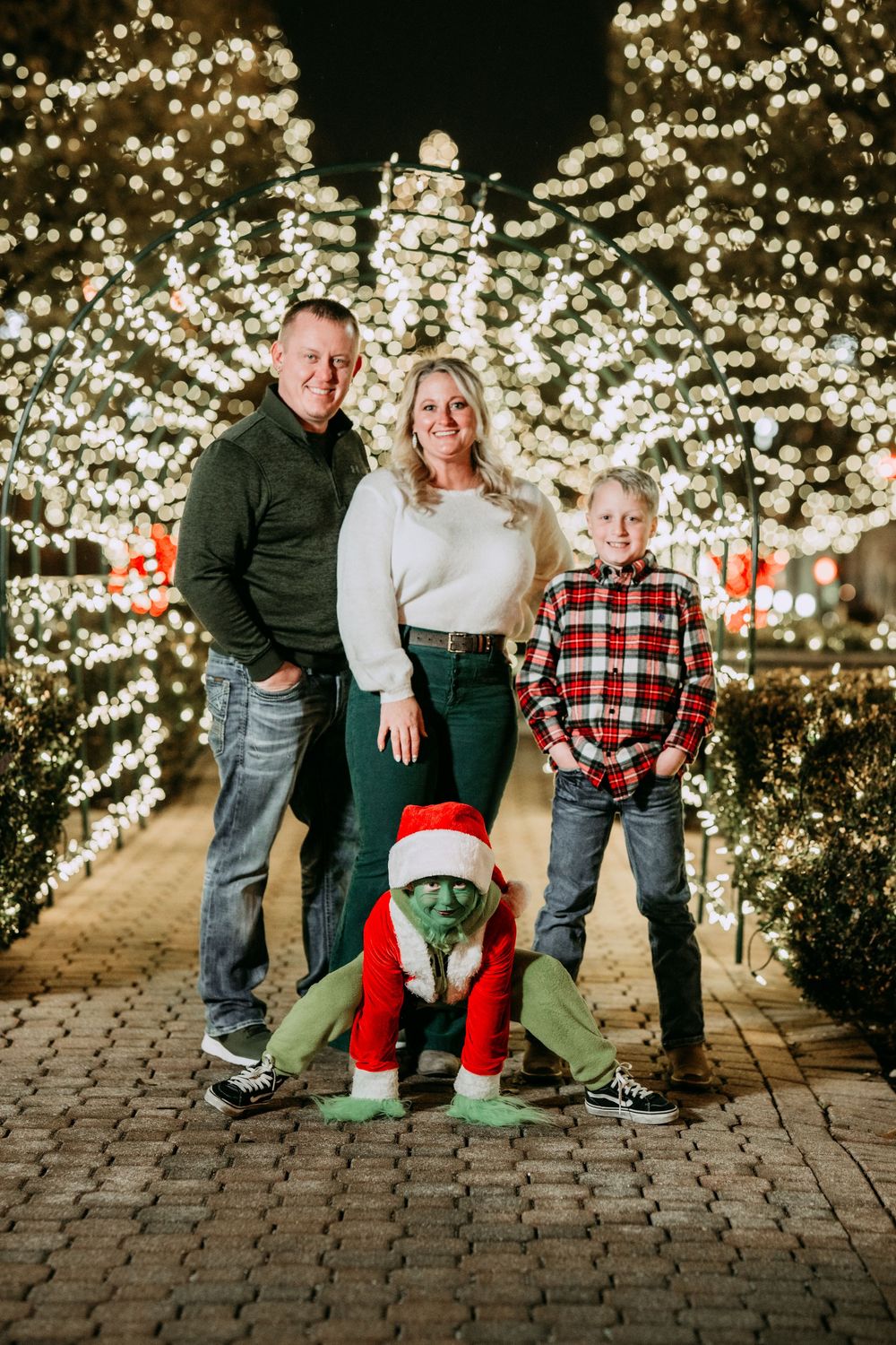 Family poses together on brick pathway surrounded by glowing Christmas trees at night.